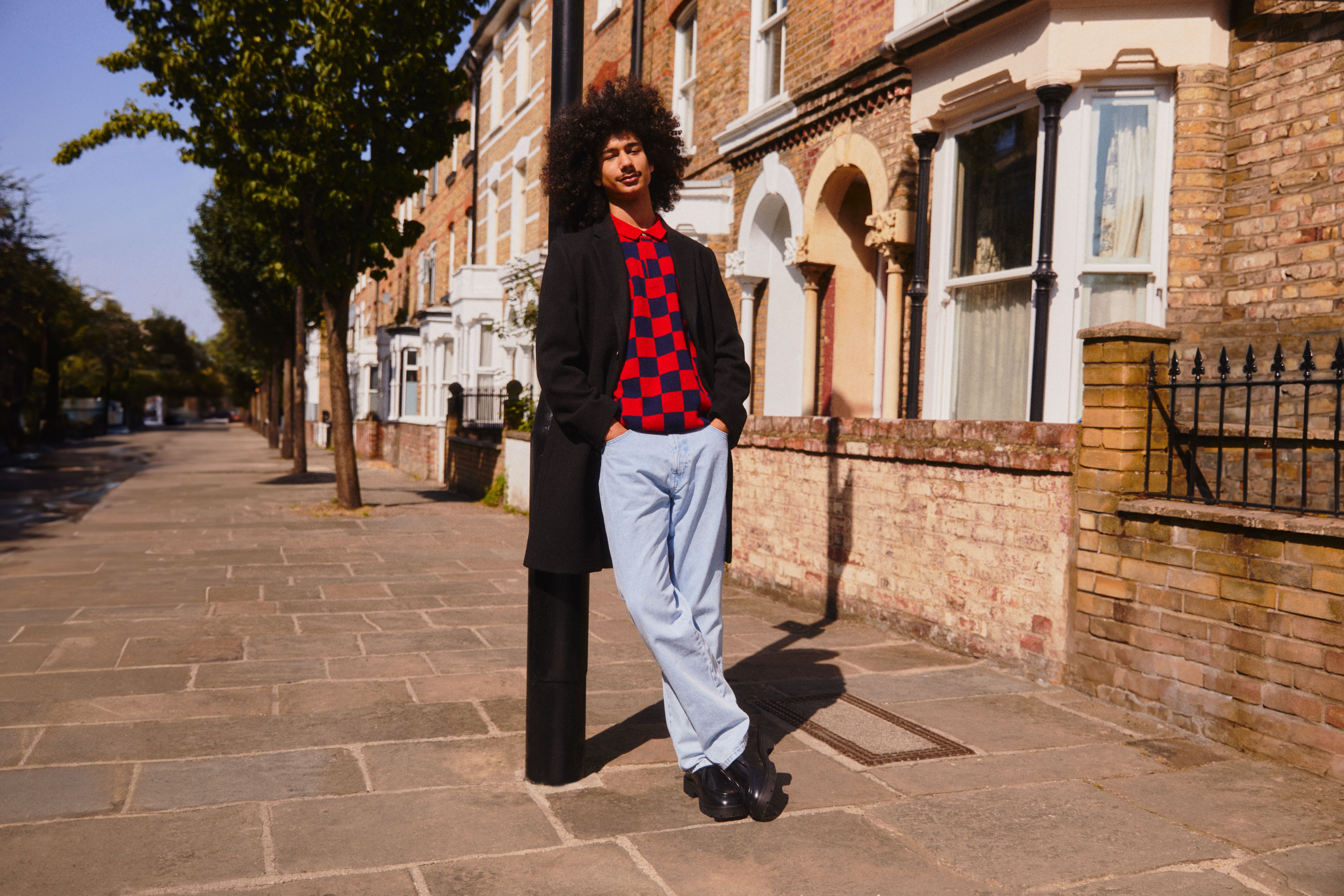 Man leunt tegen een straatlantaarn, draagt een levendige trui en lichte jeans die afsteken tegen de achtergrond